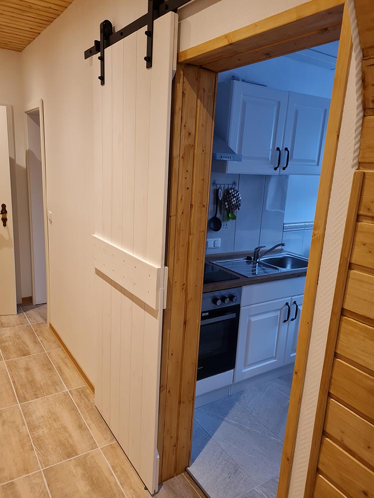 Weiß-graue Küche mit Holz-Schiebetür, hellen Wandfliesen und Edelstahl-Spüle.
