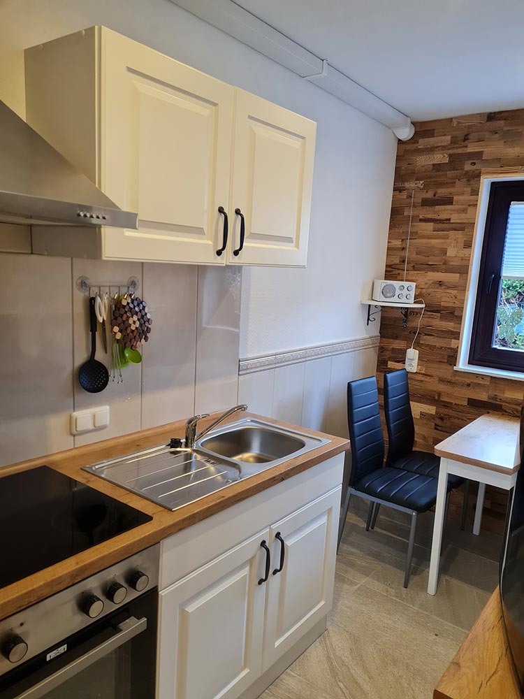 Kleine Küche mit weißen Schränken, Herd, Spüle und Tisch mit blauer Stuhl, Holzwand rechts.