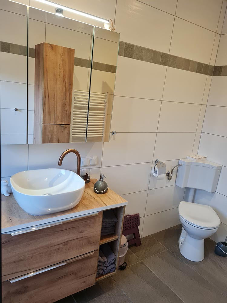 Modernes Badezimmer mit Holzschränken, rundem Waschbecken, Toilette und Wandspiegeln, weiß gefliest.