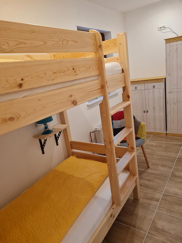 Ein heller Raum mit hölzernem Etagenbett und gelber Decke, ein bunter Stuhl im Hintergrund.