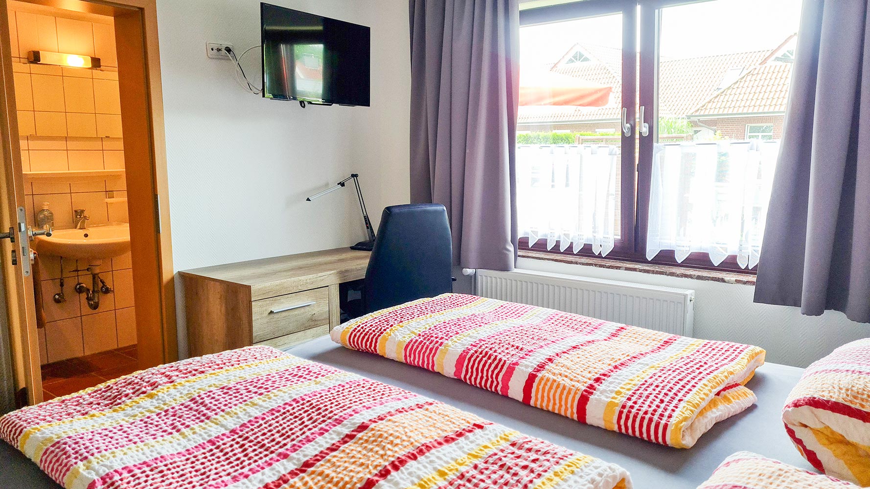 Helles Schlafzimmer mit zwei farbenfrohen Betten, einem Schreibtisch und einem Fernseher an der Wand.