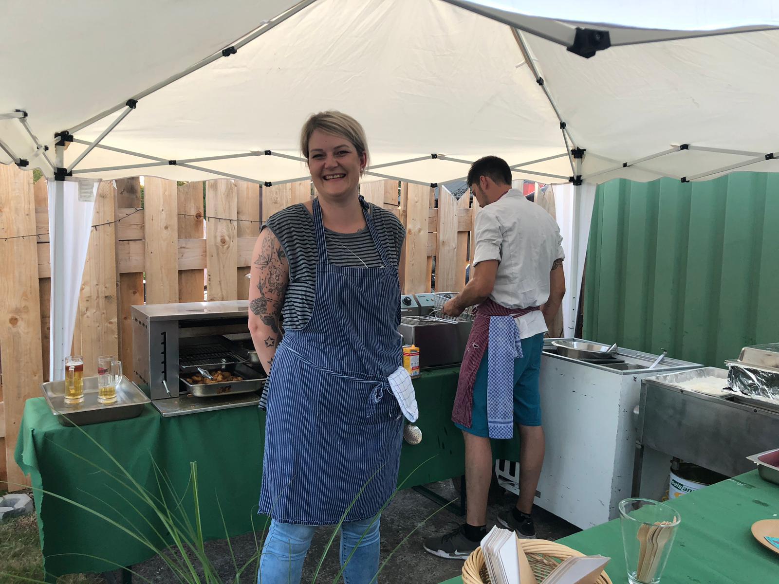 Eine lachende Frau mit Schürze und Tattoos steht draußen vor einem Grillstand unter einem Zelt.