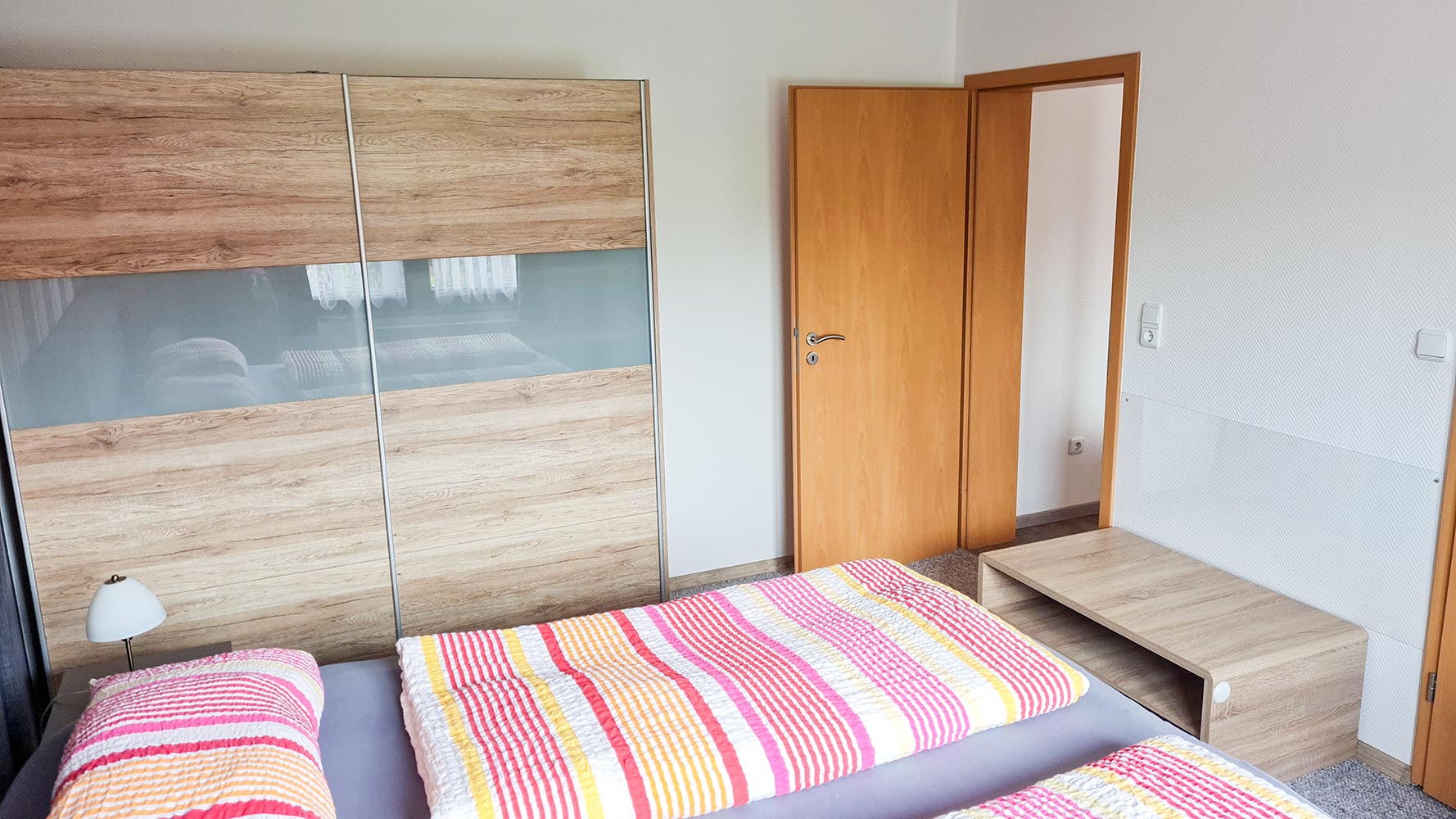 Schlafzimmer mit Holzmöbeln, bunte Bettdecken, Kleiderschrank, Nachttisch und Lampe.