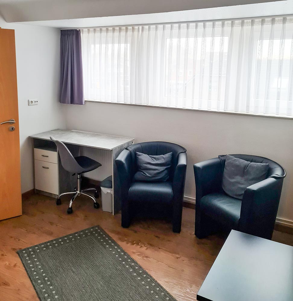 Ein kleines, einfaches Büro mit Schreibtisch, Bürostuhl, zwei schwarzen Sesseln und Tageslicht.