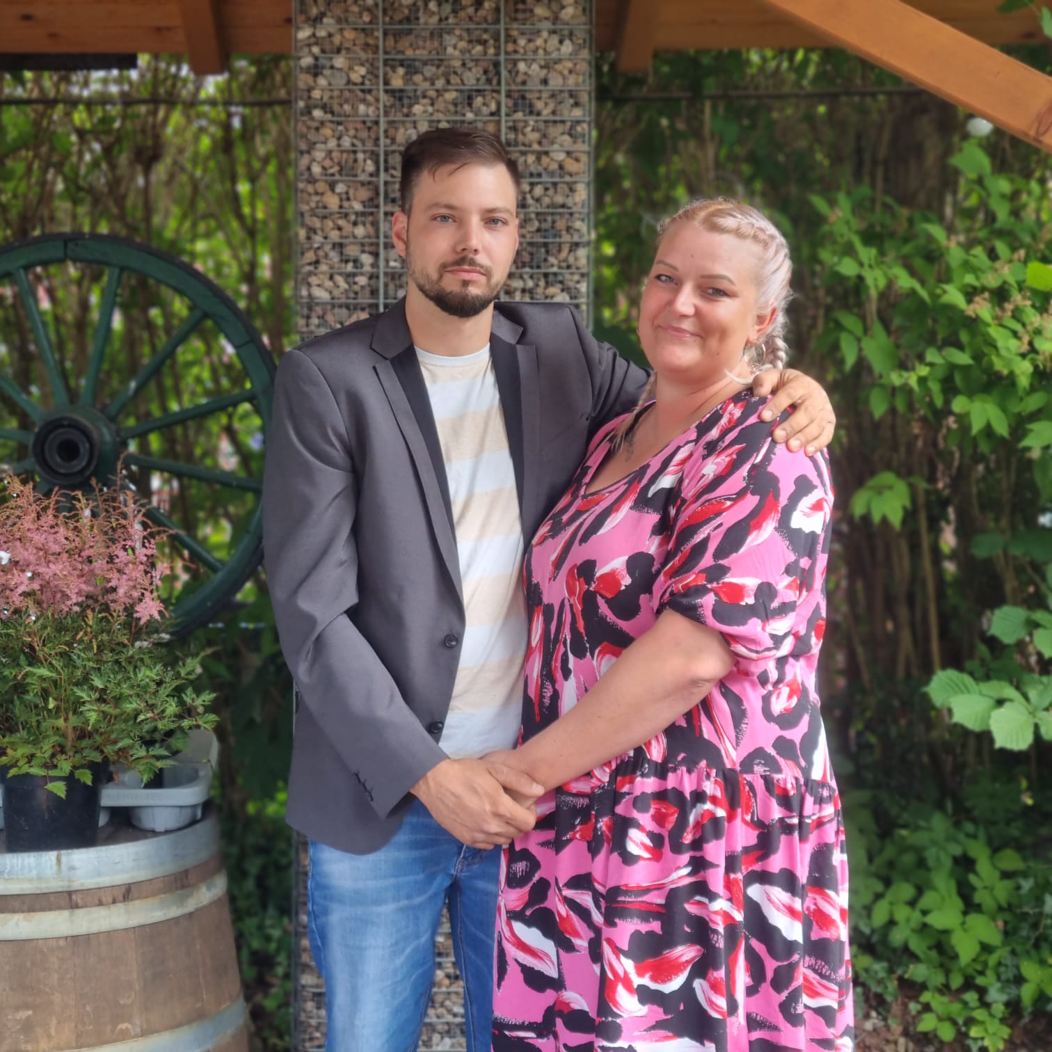 Ein Mann und eine Frau lächeln im Garten, er trägt ein Sakko, sie ein buntes Kleid.