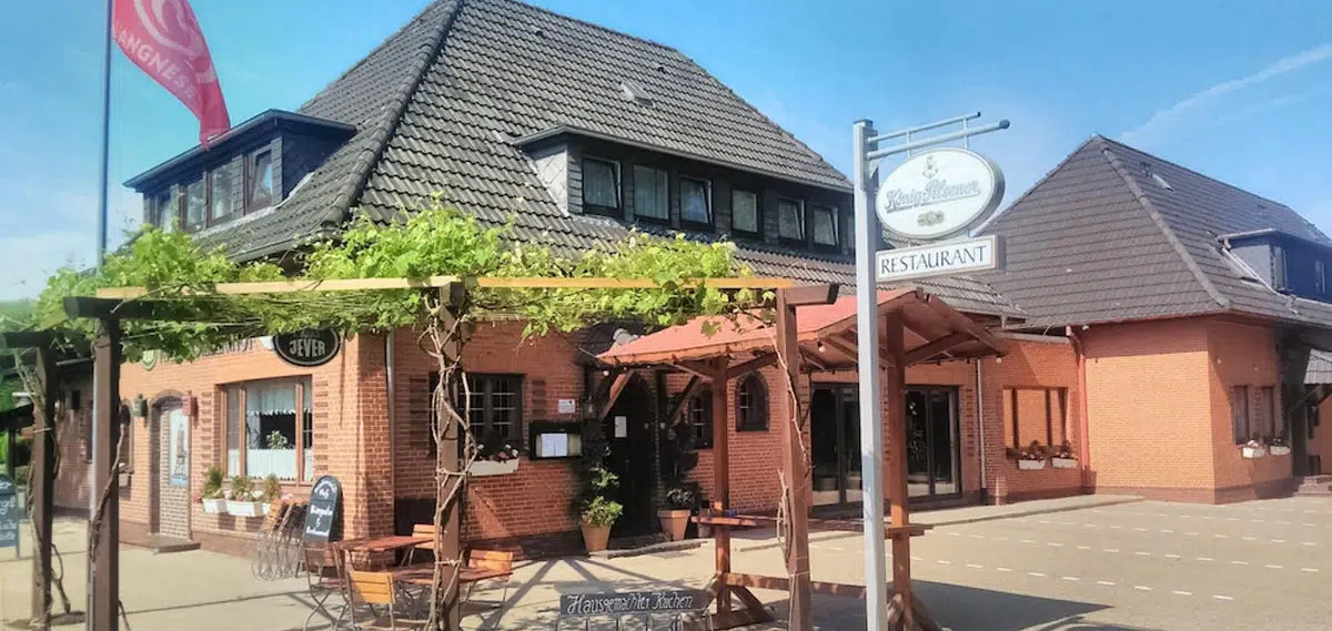 Ein rotes Backsteinrestaurant mit dunkelbraunem Schrägdach und einem Biergarten mit Pergola.
