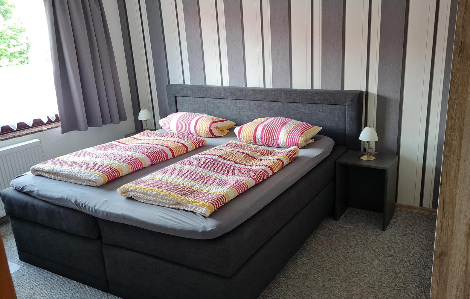 Graues Schlafzimmer mit gestreifter Tapete, Doppelbett mit bunten Decken und Lampen auf Nachttischen.