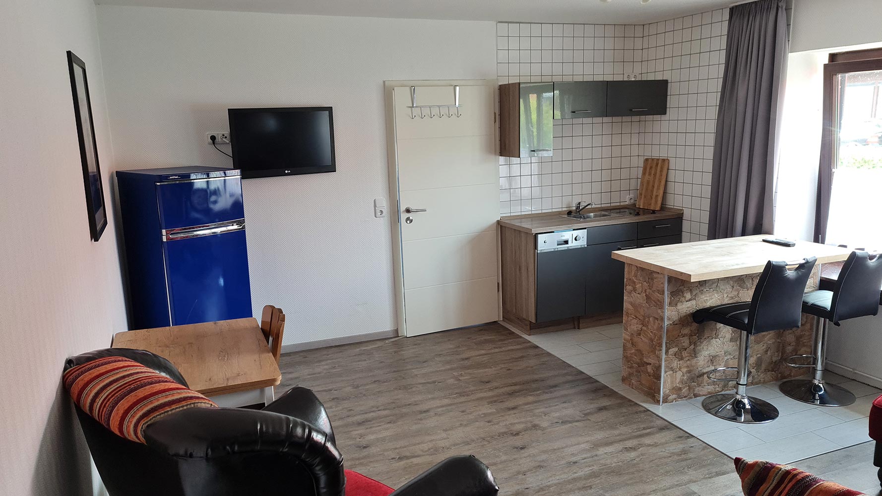 Wohnküche mit blauer Kühlschrank, Bartresen und zwei Hockern, Flachbildfernseher an der Wand.