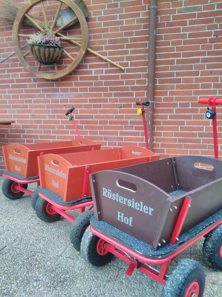 Drei rote Bollerwagen mit braunen Kisten vor einer ziegelroten Wand, dekoriert mit einem Wagenrad.
