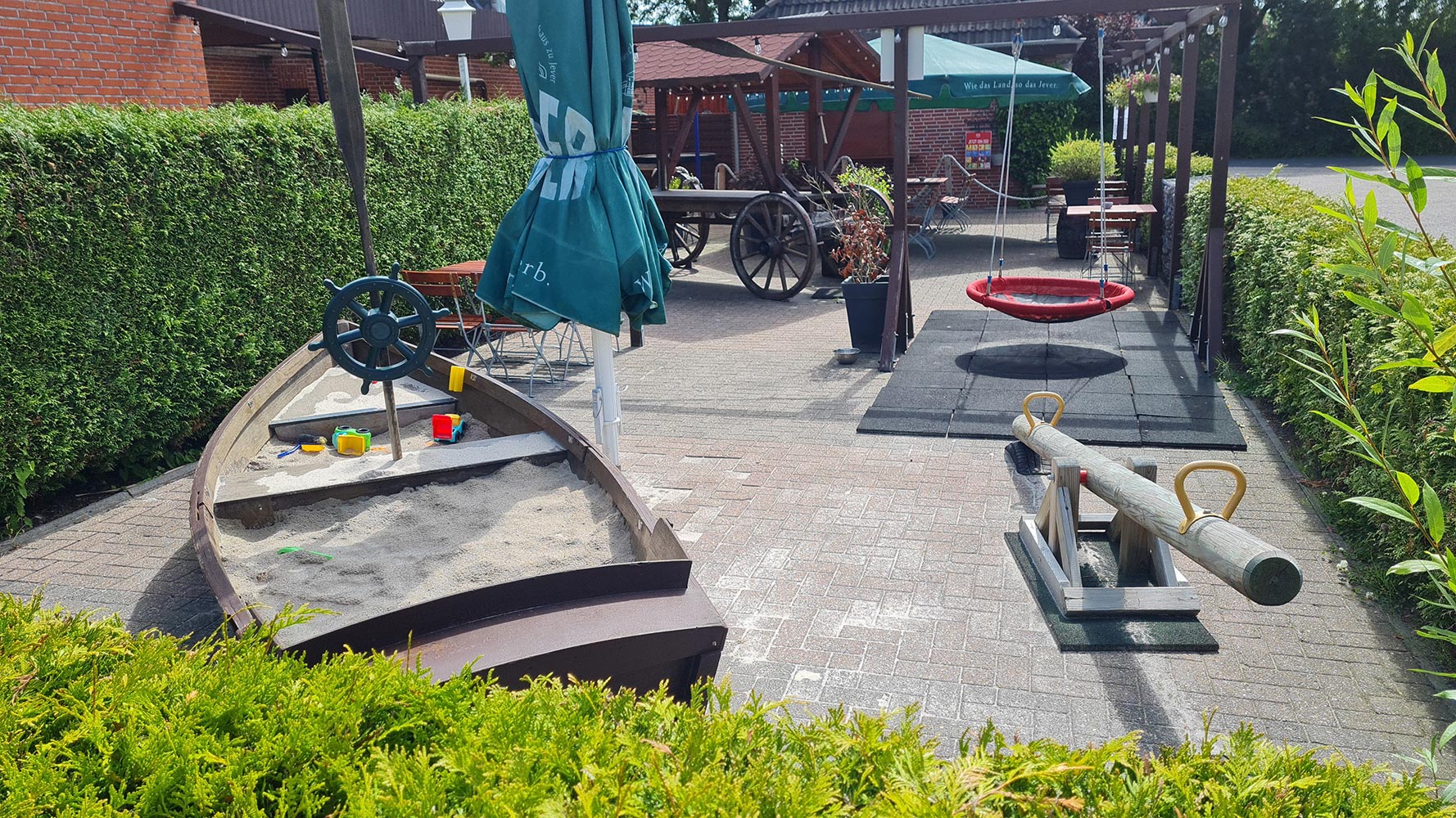 Ein Spielplatz mit einem Sandkastenschiff, Wippe und Schaukeln; umgeben von grünen Hecken.