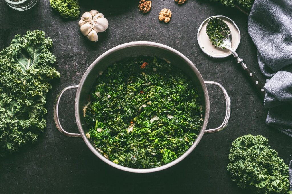 Stewed kale in cooking pot on dark kitchen table background with ingredients and spoon, top view
